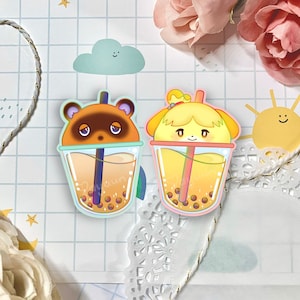 May include: Two cartoon animal characters, a raccoon and a dog, are depicted as bubble tea cups. The raccoon is brown with a light blue cup and the dog is yellow with a light pink cup. Both cups have a straw and boba pearls.