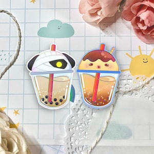 May include: Two colorful bubble tea stickers with cute cartoon faces. One sticker features a white and black panda wearing a bandage and the other sticker features a brown and white character with a chocolate topping.