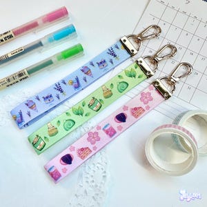 May include: Three patterned lanyards with silver clasps, featuring pastel illustrations of desserts and plants. The lanyards are light blue, green, and pink. Also visible are three pens and two rolls of washi tape.