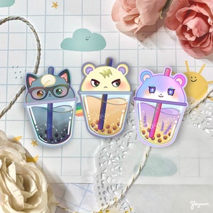 May include: Three cartoon animal characters are depicted as bubble tea cups. The characters are a cat, a hamster, and a bear. Each character has a different colored drink and boba pearls. The background is a light blue and white grid pattern.