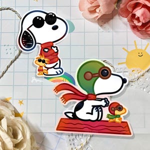 May include: Two Snoopy stickers with Woodstock. One sticker shows Snoopy wearing a red scarf and a red hat, with Woodstock. The other sticker shows Snoopy wearing a green pilot helmet and red scarf, with Woodstock.