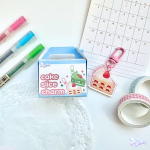May include: A light blue box with the text "cake slice charm" next to a cake slice charm on a pink keychain. The image also includes pens, washi tape, and a calendar.