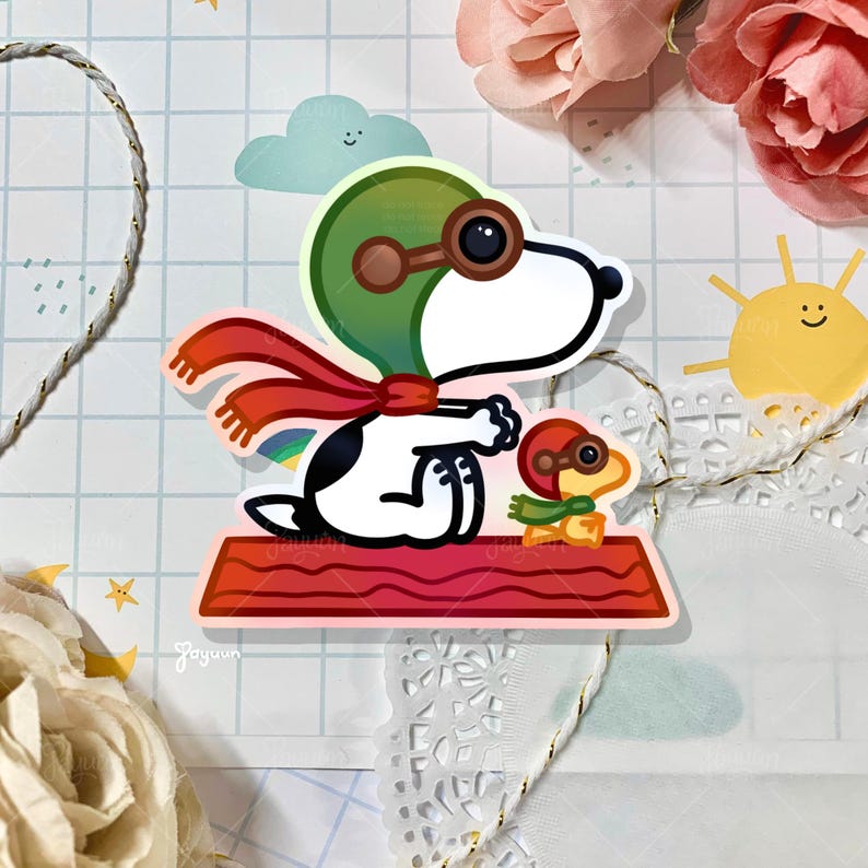 May include: A sticker featuring Snoopy as a pilot, wearing a green helmet and goggles, with a red scarf. A smaller character, also in pilot gear, is on a red platform. The background includes a rainbow, a smiling sun, and a cloud.