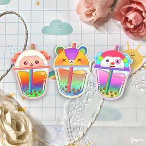 May include: Three colorful bubble tea cups with animal faces. The cups are filled with rainbow-colored liquid and boba pearls. The animals are a sheep, a bear, and a dog.