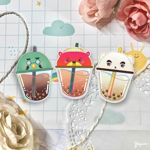 May include: Three colorful bubble tea stickers with cartoon animal faces. The stickers are on a white background with a decorative paper doily.