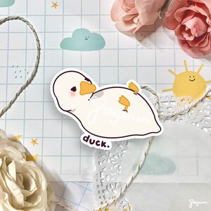 K&ouml;nnte beinhalten: Ein wei&szlig;er Aufkleber mit einer Cartoon-Ente mit orangefarbenem Schnabel und F&uuml;&szlig;en. Der Aufkleber tr&auml;gt die W&ouml;rter "Jayuun" und "duck.". Der Hintergrund umfasst ein hellblaues Gittermuster, eine Wolke und eine Sonne.