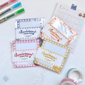 May include: A collection of memo pads with gingham patterns in various colors, including blue, orange, pink, and yellow. Each pad is labeled with a fruit theme, such as blueberry, orange, strawberry, and lemon. The pads are 3 in x 3 in. Also visible are pens, a calendar, and decorative tape.