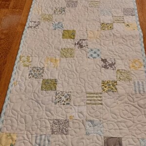 May include: A rectangular quilted table runner with a pale blue border. The runner has a patchwork design with squares in various floral and patterned fabrics, including yellow, green, and blue. The quilting pattern is a repeating floral design.