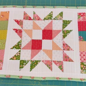 May include: A quilted table runner featuring a central star design. The star is made from red, pink, and white fabric pieces. Vertical columns of colourful squares flank the star, and a floral border frames the piece. The runner is approximately 51 cm long.