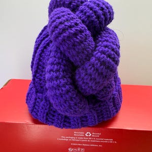 May include: A deep purple knitted hat with a textured, braided design. The hat is made of thick yarn and sits on a red box. The hat's colour is a rich, saturated purple, ideal for the colder months.