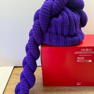 Puede incluir: Un gorro morado tejido a mano con una larga cola retorcida. El gorro es de un tono morado vibrante y presenta un patrón de puntada texturizada. La cola está asegurada con una banda elástica rosa. El gorro está sobre una caja roja.