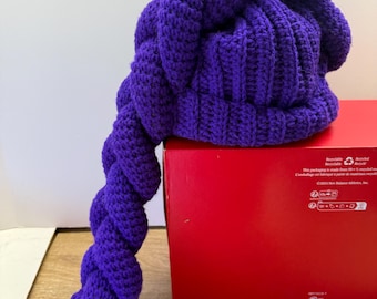 May include: A handmade, purple crochet hat with a long, twisted tail. The hat is a vibrant shade of purple and features a textured stitch pattern. The tail is secured with a pink rubber band. The hat is resting on a red box.
