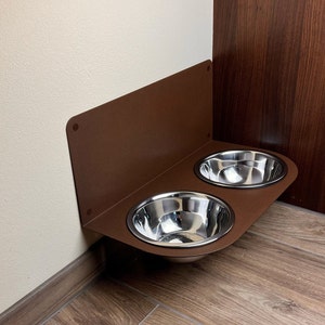 Könnte beinhalten: Ein brauner Metall-Futternapfhalter, der an einer Wand montiert ist, mit zwei Edelstahlnäpfen.