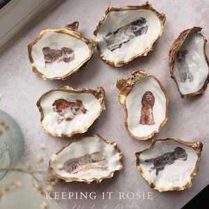 May include: Seven decorative oyster shells with gold-painted edges and hand-painted dog illustrations on the interior. The text KEEPING IT ROSIE Handmade Gifts is centred at the bottom. Each shell features a different dog breed in various poses.