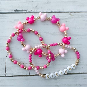 May include: Four pink and gold beaded bracelets with Mickey Mouse head charms. One bracelet has the word "SISTERS" spelled out in white beads.