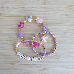 May include: A set of three beaded bracelets with gold accents. The bracelets feature purple, pink, and light blue beads, as well as small Mickey Mouse charms. One bracelet has the name "MADISON" spelled out in white letters.