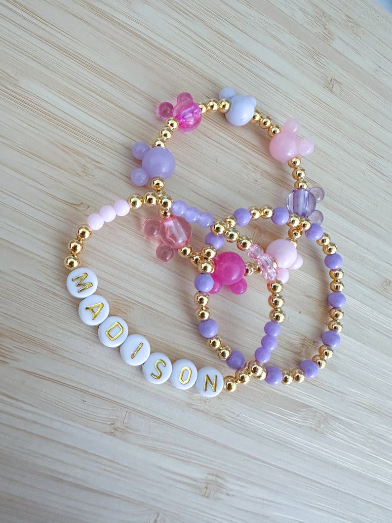May include: Three bracelets with gold beads and colorful charms. One bracelet has the name "MADISON" spelled out in white beads with gold lettering.