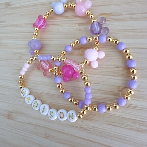 May include: A set of three bracelets with gold beads and colorful charms. The bracelets feature a name spelled out in white beads with gold lettering, "MADISON". The charms include pink, purple, and white Mickey Mouse ears.