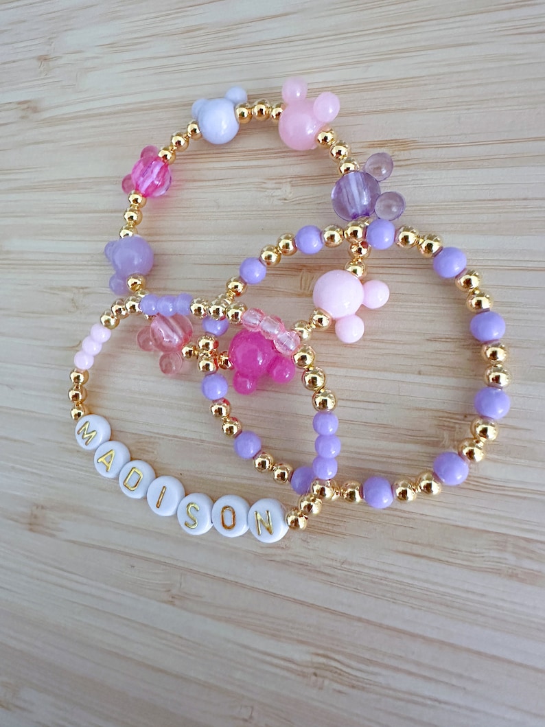 May include: A set of three bracelets with gold beads and colorful charms. The bracelets feature a name spelled out in white beads with gold lettering, "MADISON". The charms include pink, purple, and white Mickey Mouse ears.