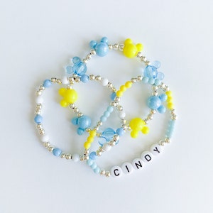 Op de afbeelding: Drie armbanden van kralen in blauw, geel en wit. De armbanden hebben Mickey Mouse-vormige kralen en zilveren accenten. E&eacute;n armband spelt de naam "CINDY" met witte letterkralen.