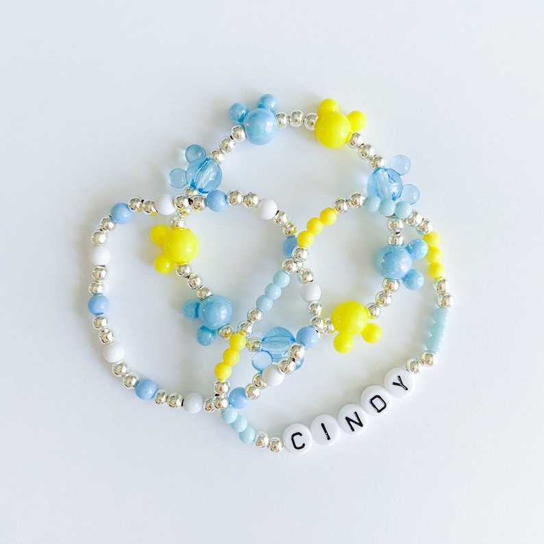 Op de afbeelding: Drie armbanden van kralen in blauw, geel en wit. De armbanden hebben Mickey Mouse-vormige kralen en zilveren accenten. E&eacute;n armband spelt de naam "CINDY" met witte letterkralen.