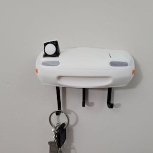 Puede incluir: Porta llaves blanco en forma de coche con tres ganchos y una pequeña luz negra en la parte superior. Una llave cuelga de uno de los ganchos.