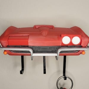 Könnte beinhalten: Ein roter, auto-förmiger Schlüsselhalter mit Chromstoßstange und schwarzen Schlüsselhaken. Der Schlüsselhalter hat zwei weiße Scheinwerfer und orangefarbene Seitenmarkierungen. Das Design ähnelt der Front eines klassischen Autos.