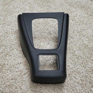 May include: A black plastic automotive console trim piece. The trim has a rectangular opening at the top and a smaller square opening at the bottom. The piece is designed to fit around the gear shift and other controls.