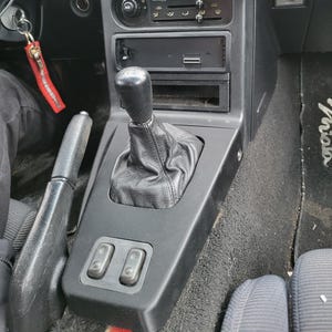 May include: Black leather gear shift boot with a black plastic gear shift lever. The gear shift is in a car's centre console with black plastic buttons.