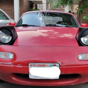 Peut inclure: Une Mazda Miata rouge avec des phares noirs et une plaque d'immatriculation du Wisconsin. La voiture est garée sur un parking.