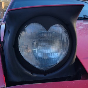 May include: A red car with a heart-shaped headlight. The headlight is made of clear glass and has a black plastic frame.
