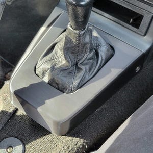 Op de afbeelding: Een zwarte leren versnellingspookhoes met een zwarte kunststof versnellingspookknop in het interieur van een auto. De versnellingspook is omgeven door een grijze kunststof console.