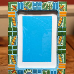 May include: A rectangular picture frame with a mosaic tile design. The frame features a blue border with green, orange, and blue mosaic tiles. The frame holds a blue background, ready for a photo.