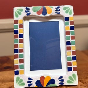 May include: A rectangular photo frame with a white interior and a colorful mosaic tile border. The border features small square tiles in shades of red, green, yellow, and blue. The top and bottom of the frame have decorative floral designs in various colors.