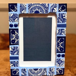 May include: A rectangular picture frame decorated with a mosaic of blue and white ceramic tiles featuring floral and geometric patterns. The tiles are set in light blue grout, and the frame surrounds a dark blue rectangular insert.