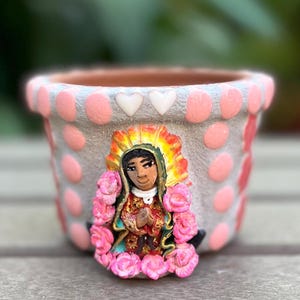May include: A small, decorative terracotta pot with a gray base, adorned with pink polka dots and a sculpted image of a religious figure. The figure is surrounded by pink roses and has a gold halo. Two white heart-shaped accents are near the rim.