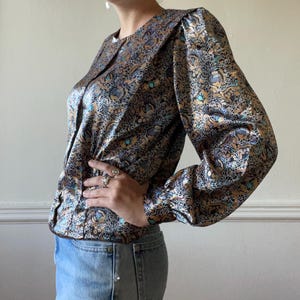 May include: A long-sleeved blouse featuring a metallic gold, black, and light blue intricate floral pattern. The garment has a round neckline, a button-down front, and gathered sleeves with cuffs, paired with light blue denim jeans.