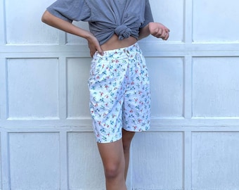 May include: High-waisted white shorts featuring a delicate floral pattern with small red and blue flowers and green leaves. The shorts have a button closure at the waist, a pleated front design, and a tailored fit that reaches mid-thigh.