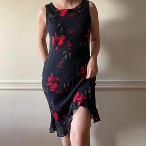 May include: Sleeveless black dress with a red floral pattern. The dress has a knee-length hemline and a scoop neckline. The red flowers are scattered across the dress, with green leaves. The dress is made of a lightweight fabric.