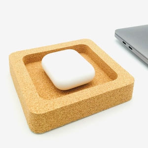 Peut inclure: Un étui de charge sans fil blanc rectangulaire est placé dans un plateau carré en liège. Le plateau est de couleur brun naturel et a une surface texturée.