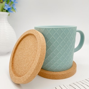 Könnte beinhalten: Eine hellblaue Keramiktasse mit einem Rautenmuster steht auf einem Korkuntersetzer. Ein zweiter Korkuntersetzer befindet sich neben der Tasse.