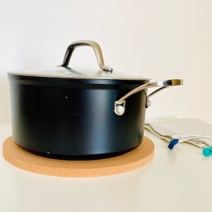 May include: A black pot with a glass lid and silver handles sits on a round cork trivet.