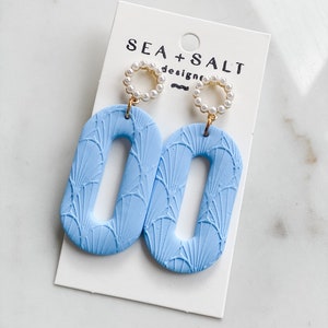 May include: A pair of light blue ceramic earrings with a fan-shaped pattern. The earrings are oval-shaped with a hole in the center. The earrings are attached to a gold-colored post with a pearl circle.
