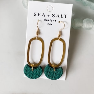 May include: Gold oval earrings with a green leaf design dangle from the bottom. The earrings are on a white card with the text "SEA + SALT designs".