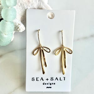 May include: Gold bow earrings with a simple design. The earrings are on a white card with the text "SEA + SALT designs".