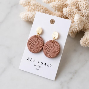 May include: A pair of circular earrings featuring a dusty rose textured pattern of palm-like leaves, suspended from small gold-toned hammered metal discs. The earrings are attached to a white display card printed with the text SEA + SALT designs.