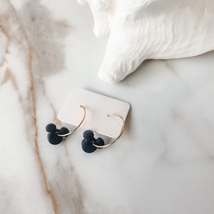 May include: Black Mickey Mouse-shaped earrings with gold-coloured hoops. The earrings are presented on a white card against a marble background, with a white seashell in the background.