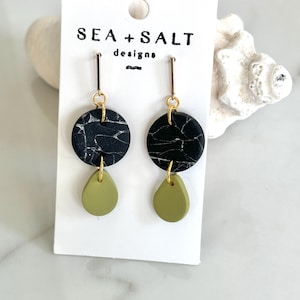 Puede incluir: Un par de pendientes colgantes con tapas redondas de mármol blanco y negro y fondos de lágrima verde. Los pendientes están en una tarjeta blanca con el texto "SEA + SALT designs".