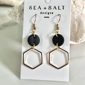 Könnte beinhalten: Ein Paar schwarz-goldene Ohrringe mit geometrischem Design. Jeder Ohrring hat einen schwarzen runden Ohrstecker, der mit einer goldenen Sechseckform verbunden ist. Die Ohrringe werden auf einer weißen Karte mit dem Text "SEA + SALT designs" präsentiert.