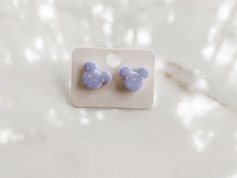 May include: A pair of lavender-colored stud earrings shaped like a cartoon mouse head with white polka dots. The earrings are displayed on a white card with a hole at the top.